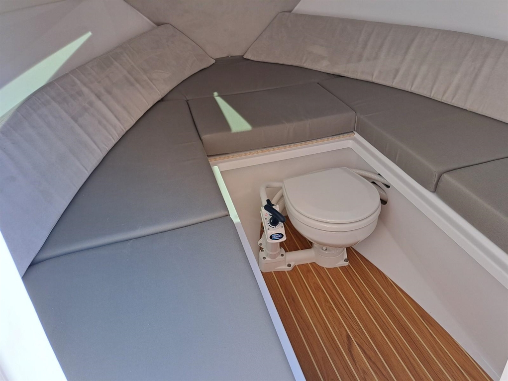 Interior of a boat featuring gray cushioned seating, a wooden floor, and a compact marine toilet in a well-lit space.